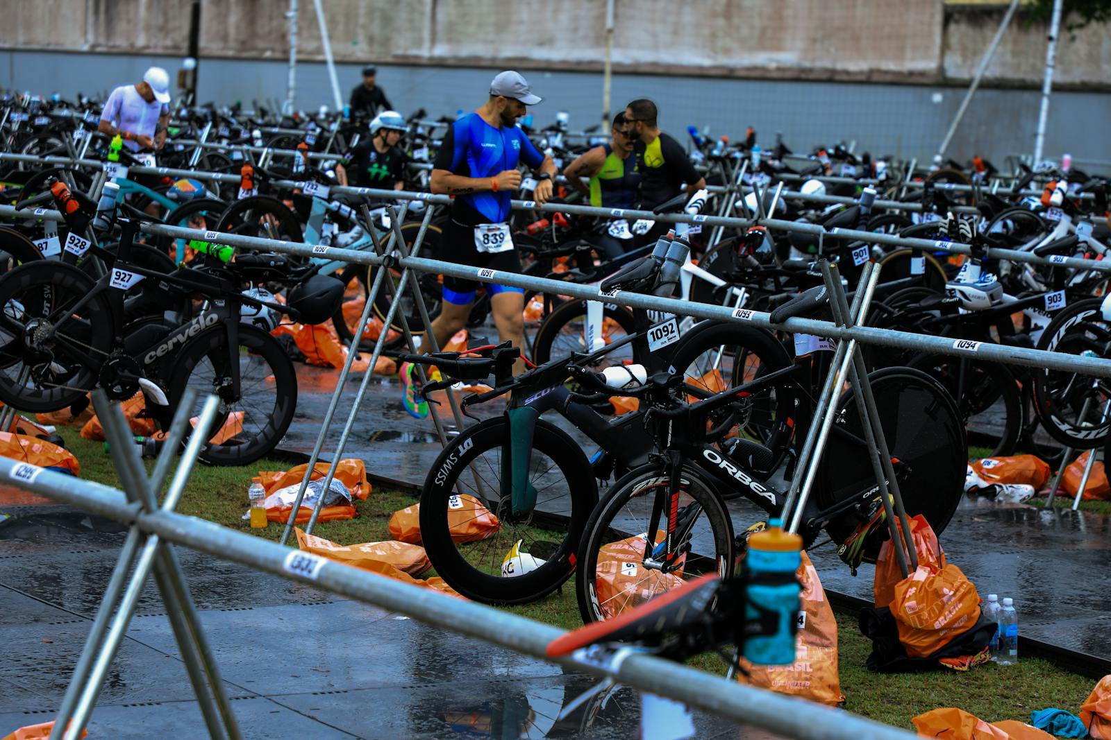 Athletes in a triathlon transition zone surrounded by multiple bicycles and gear.